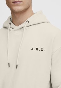 Sweat à capuche beige en tissu doux, avec une poche avant, une capuche à cordon de serrage, et un logo noir "A.R.C." sur le côté gauche de la poitrine.