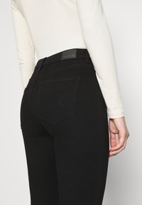 Jean en denim noir à taille haute, avec des poches arrière et un écusson en cuir avec le logo à la ceinture. Texture lisse et coupe ajustée.