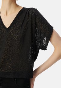 Top de estampado de leopardo negro hecho de tela transparente. Presenta un escote en V, mangas cortas y un corte suelto. La textura sutil añade profundidad.