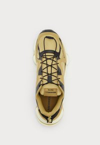Zapatilla deportiva marrón con parte superior de malla, acentos negros y sistema de cordones integrado. Diseño de punta redondeada y suela acolchada.