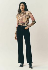 Kobieta stoi ubrana w długi rękaw kwiecisty top, czarne spodnie z wysokim stanem oraz czarne botki, trzymając jedną rękę w kieszeni.