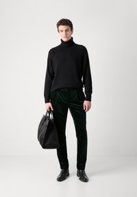 Black turtleneck sweater, green velvet pants, black leather shoes, and a large black bag. Smooth texture and fitted design throughout.