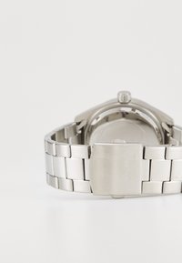 Stříbrný kovový řetězový pásek hodinek se sponou s rytinou "GUESS", zobrazený zezadu s ciferníkem hodinek mimo záběr na bílém podkladu.