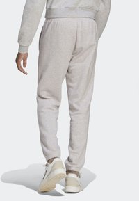 Person som bär ljusgrå prickiga sweatpants och beigevita sneakers, sedd bakifrån när de går på en enfärgad ljus bakgrund.