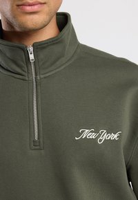 Sudadera con cremallera de color verde oscuro y cuello alto, con una cremallera plateada y texto bordado en blanco que dice "New York" en el pecho.