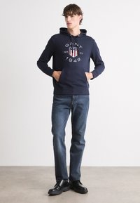 Sudadera con capucha azul marino y bolsillo frontal, con un gráfico blanco que presenta un diseño de escudo rojo y azul, combinada con jeans azules y zapatos negros.