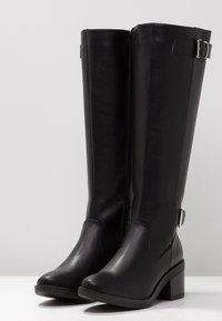 Para czarnych skórzanych botków do połowy łydki z blokowym obcasem, zamkami błyskawicznymi po bokach i ozdobnymi srebrnymi klamrami w pobliżu góry i kostki.