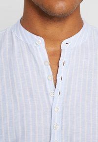 Chemise en lin bleu clair à rayures verticales avec un col mandarin et sept boutons blancs. Présente un col partiellement déboutonné avec un design décontracté.