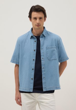 Junger Mann mit dunklem Haar, der ein hellblaues kurzärmeliges Jeanshemd über einem schwarzen T-Shirt und weißen Hosen trägt, vor hellem Hintergrund stehend.