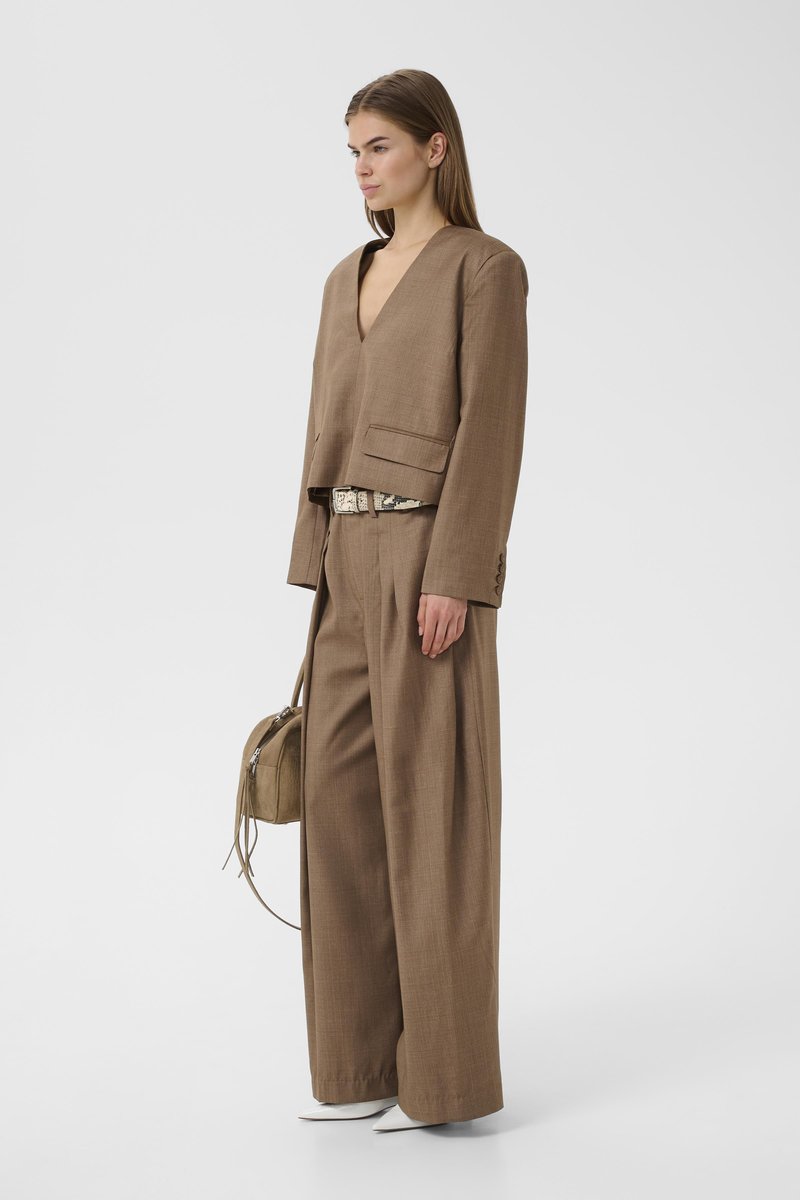 Femme portant un costume ample marron avec un pantalon à jambes larges, une ceinture en peau de serpent, des chaussures blanches à bouts pointus et un sac à main marron assorti.