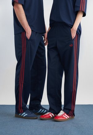 To personer iført marineblå treningsbukser og skjorter med røde striper, som står side om side med hendene i lommene.