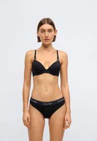 Must pehmendusega rinnahoidja õhukeste rihmadega, sileda tekstuuriga ja mustadel sobivatest püksikutest koosneval komplektil on logoga pael. Lihtne disain, minimalistlik stiil.