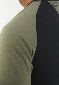 Kortärmad skjorta med en olivgrön kropp och svarta raglanärmar. Tyget verkar mjukt med en slät textur och subtila sömnadsdetaljer.