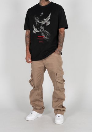 Black t-shirt with a graphic featuring chains and two doves, paired with beige cargo pants featuring multiple pockets and white sneakers.