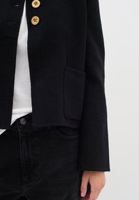 Černý vlněný blazer s třemi velkými béžovými knoflíky, přední kapsou a boxy střihem. Spárován s černými džínami a bílým tričkem pod tím.