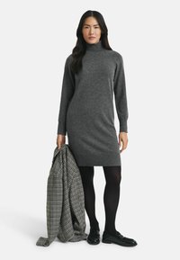 Abito grigio lavorato a maglia con collo alto, maniche lunghe e orlo a coste. Il modello tiene in mano un cappotto a stampa bianca e nera e indossa collant e scarpe nere.
