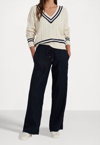 Krémový svetr s copánkovým vzorem a námořnicky modrými pruhy na výstřihu do V a na manžetách, doplněný volnými tmavě modrými kalhotami s kapsami.