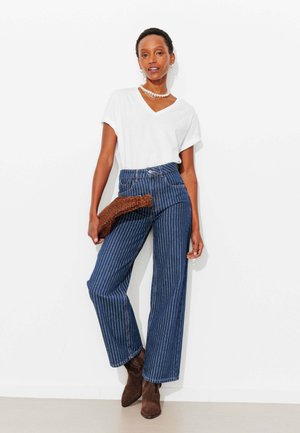 Femme portant un t-shirt blanc à col en V, un jean large à rayures fines bleu taille haute, des bottes en suède marron, tenant une pochette à motif léopard.
