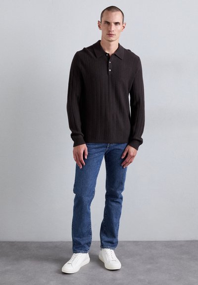 Temno rjav pleten polo pulover z rebrasto teksturo, ki ima klasičen ovratnik in tri gumbe, v kombinaciji z modrimi denim hlačami in belimi športnimi copati.