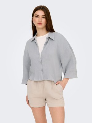 Vrouw draagt lichtgrijs overhemd met knopen en brede mouwen over een wit topje, gecombineerd met beige korte broek, staand met één hand in de zak.