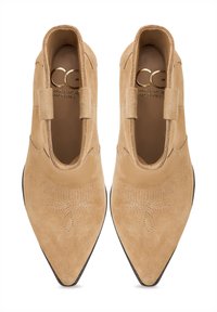 Botine scurte din suede bej, cu vârf ascuțit, panouri laterale elastice și cusături decorative. Textură netedă și detalii hardware minime.