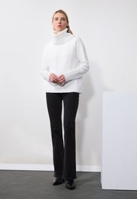 Maglione bianco a collo alto lavorato a maglia con texture a coste, abbinato a pantaloni neri a zampa e stivaletti neri. Design semplice, senza motivi visibili.