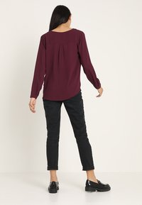 Blusa bordeaux a maniche lunghe con scollatura tonda e dettaglio a pieghe sul retro, abbinata a jeans neri a gamba dritta e scarpe in pelle nere con plateau.