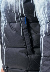 Primer plano de una chaqueta acolchada negra y gris con patrones de rayas blancas y un bolsillo vertical con cremallera y tirador azul.