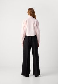 Camicia rosa cropped con maniche lunghe, chiusura a bottoni sul retro. Abbinata a pantaloni neri a gamba larga, creando un contrasto di colore e forma.