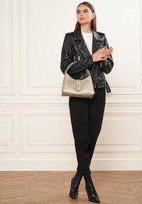 Giacca da biker in pelle nera sopra una camicia bianca, pantaloni neri e stivaletti neri a punta. Tiene in mano una borsa metallica dorata con chiusura.