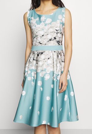 Een mouwloze teal jurk met een aangesloten lijfje en een uitlopende rok, met een bloemmotief van witte bloemblaadjes en een bijpassende riem in de taille.