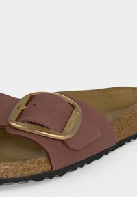 Κοντινή λήψη καφέ δερμάτινου σανδάλιου με μεγάλη χρυσή πόρπη, φελλό σόλα και μαύρο ανάγλυφο πάτο.