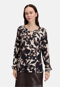Long-sleeve blouse in a cream base with brown and black floral print. Features a v-neckline with a chain accent and gathered sleeves.