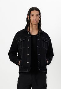 Schwarze Denimjacke mit kontrastierenden weißen Nähten, Knopfverschluss, zwei Vorderaschen und spitzem Kragen. Glatte Textur.