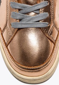 Sneaker metallico bronzo con lacci grigi, caratterizzato da una finitura liscia, forma arrotondata e suola in contrasto color nocciola. Cuciture dettagliate visibili.