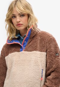 Pull en polaire marron et beige avec un col, ornée d'accents bleu et rouge, mise en valeur par un écusson logo et des textures contrastantes.