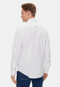 Camicia bianca a maniche lunghe con colletto a punta, tessuto liscio e polsini con bottoni. La schiena presenta un yoke e una leggera curvatura all'orlo.