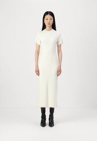 Mekko, vaaleanbeige neule midi-pituudella, jossa lyhyet hihat, sivuaukot ja muotoonommeltu siluetti; yhdistettynä mustiin, polviin ylettyviin, teksturoituihin saappaisiin.