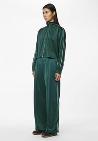 Mørkegrøn atletisk træningsdragt lavet af glat stof. Har en jakke med lynlås, afslappede bukser og kontrasterende hvide sidestriber.