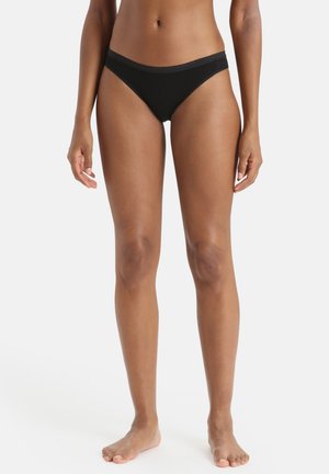 Frau steht barfuß und trägt schwarze Bikini-Unterwäsche vor einem schlichten weißen Hintergrund.