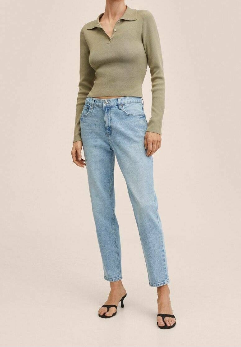 Femme portant un polo court à manches longues vert olive, un jean taille haute bleu clair et des escarpins ouverts noirs à talon kitten, debout devant un fond beige.