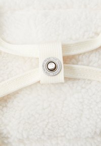 Hvit fleecestoff med en teksturert overflate, prydet med en kremfarget stropp som holder en sølvmetallknapp med preget merkevare.