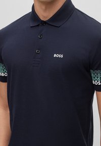 Polo bleu marine en tissu respirant ; doté d'un col à deux boutons et de manches courtes ornées de liserés avec le logo "BOSS" répété.