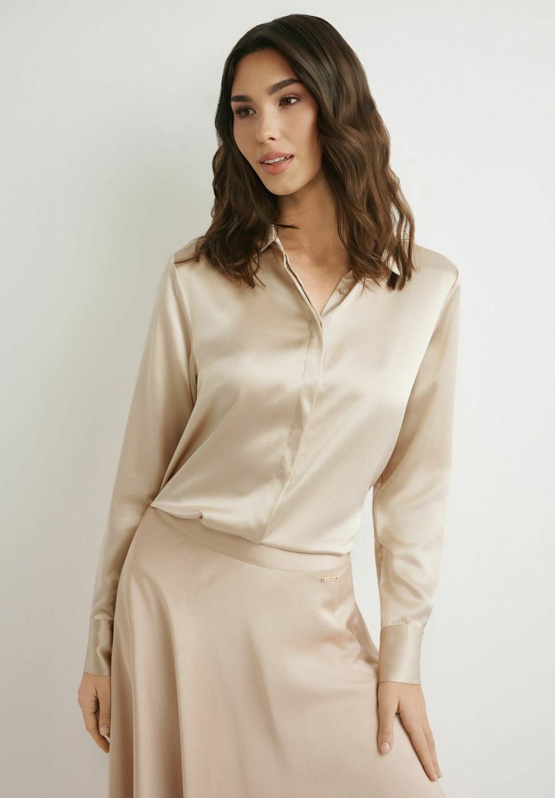Kobieta o średniej długości falowanych ciemnych włosach, ubrana w beżową satynową koszulę z długim rękawem oraz pasującą spódnicę, stojąca na tle jednolitego tła.