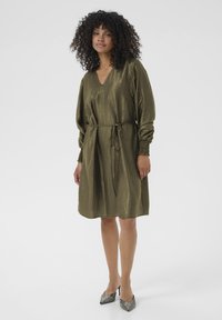 Femme aux cheveux bouclés portant une robe olive longue jusqu'aux genoux avec des manches longues et une ceinture à la taille, se tenant debout dans des chaussures à talons hauts scintillantes.
