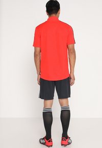 Mies seisoo selin, päällään punainen urheilupaita, mustat shortsit, mustat polvenmittaiset sukat valkoisilla raidoilla ja punaiset nappulakengät.