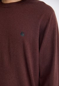 Bordeauxfarbener Strickpullover mit Rundhalsausschnitt, der ein dezentes schwarzes Logo auf der linken Brust zeigt. Weicher Stoff und figurbetonte Silhouette.