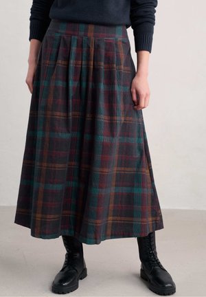Donna che indossa una gonna midi plissettata a quadri scuri, maglione blu navy e stivali neri stringati, in piedi davanti a uno sfondo semplice.