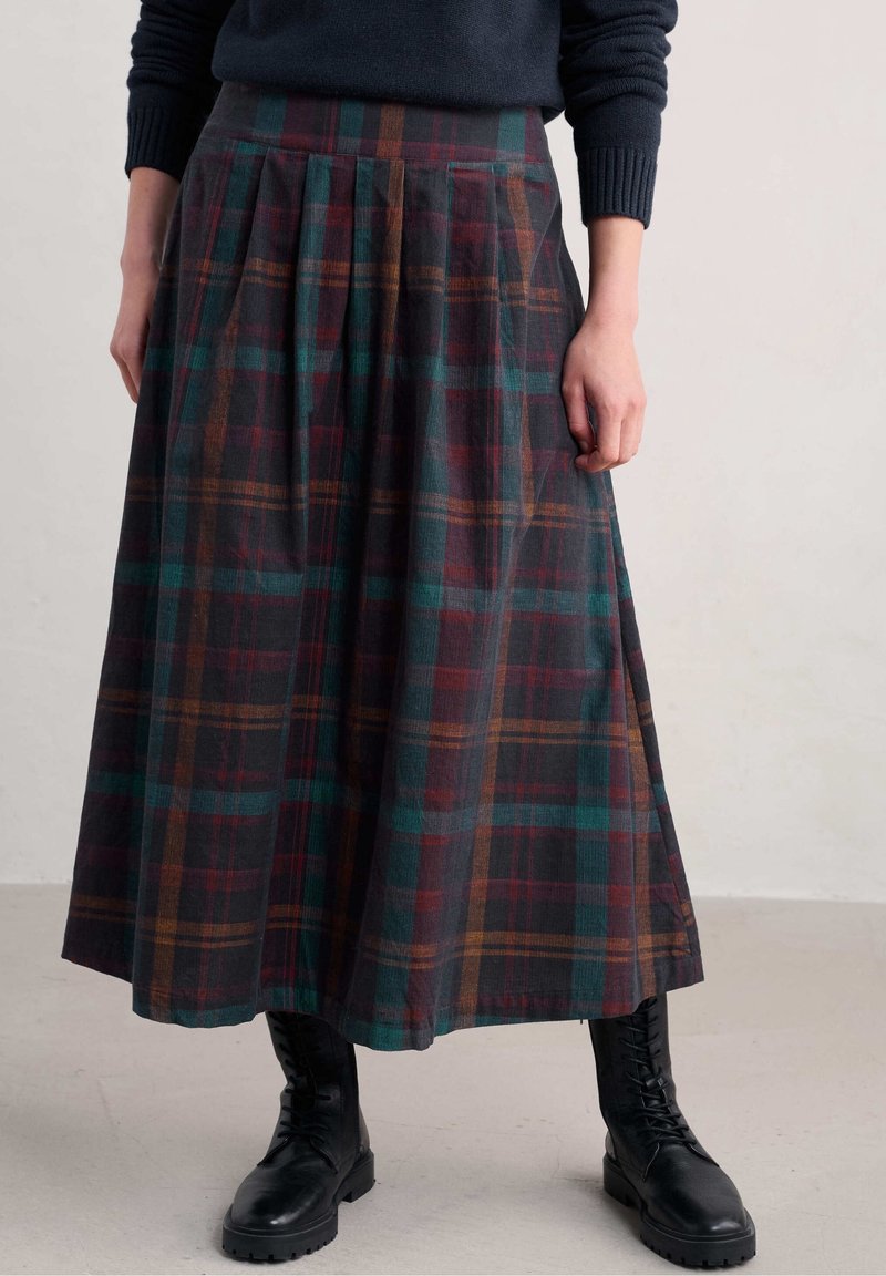 Femme portant une jupe midi plissée à carreaux foncés, un pull bleu marine et des bottes noires à lacets, debout devant un fond uni.