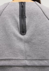 Grå sweatshirt med dragkedja baktill och svarta detaljer samt en mjuk textur. Den övre delen har en rund halsringning med sömdetaljer.